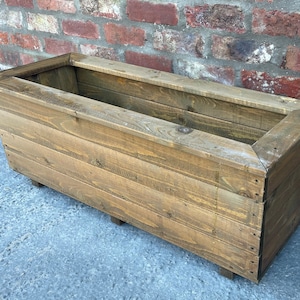 May include: A rectangular wooden planter box with a natural brown finish. The planter is made of reclaimed wood and has a rustic look.