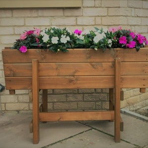 May include: A wooden planter box with a brown stain finish, sitting on a wooden stand. The planter is filled with pink and white flowers.