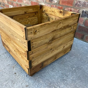 May include: A square wooden planter box with a natural wood finish. The planter is made of multiple pieces of wood that are joined together to create a sturdy and durable planter.