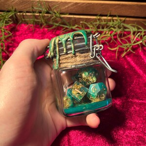 Pode incluir: Um pequeno frasco de vidro cheio de dados verde-azulado e dourados. O frasco tem um fecho de metal e &eacute; coberto por uma tampa decorada com tinta verde e dourada. Os dados s&atilde;o vis&iacute;veis atrav&eacute;s do vidro transparente.