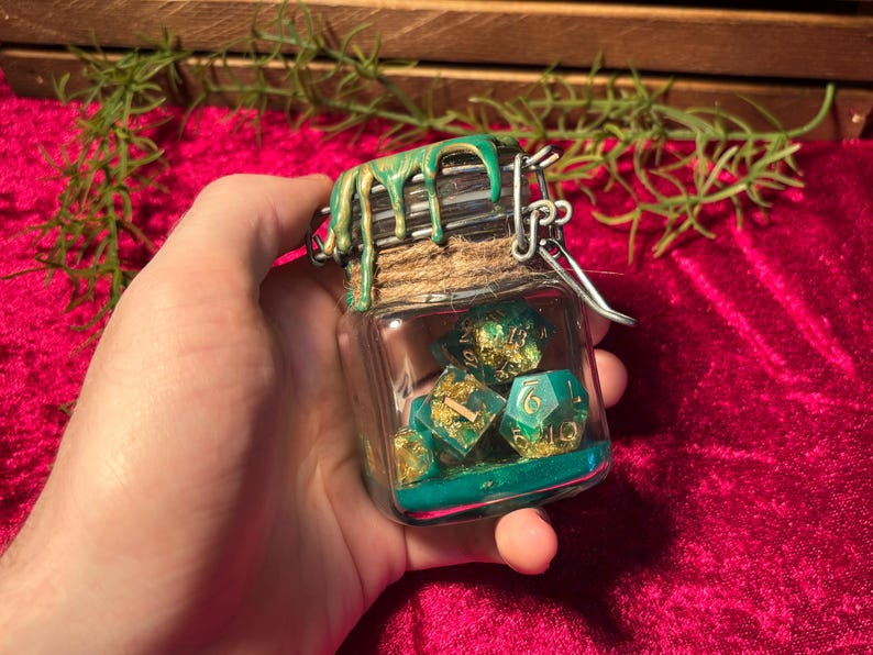 Pode incluir: Um pequeno frasco de vidro cheio de dados verde-azulado e dourados. O frasco tem um fecho de metal e &eacute; coberto por uma tampa decorada com tinta verde e dourada. Os dados s&atilde;o vis&iacute;veis atrav&eacute;s do vidro transparente.