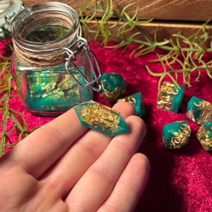 Pode incluir: Um conjunto de dados de resina feitos &agrave; m&atilde;o em tons de verde-azulado e dourado. Os dados s&atilde;o exibidos em uma superf&iacute;cie vermelha, com um frasco de vidro e vegeta&ccedil;&atilde;o ao fundo. Um dado &eacute; segurado em uma m&atilde;o.