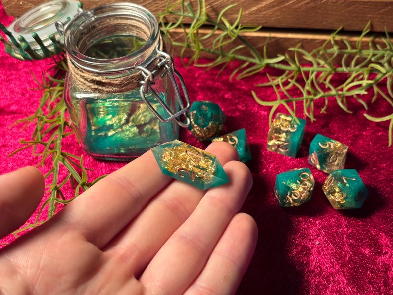 Pode incluir: Um conjunto de dados de resina feitos &agrave; m&atilde;o em tons de verde-azulado e dourado. Os dados s&atilde;o exibidos em uma superf&iacute;cie vermelha, com um frasco de vidro e vegeta&ccedil;&atilde;o ao fundo. Um dado &eacute; segurado em uma m&atilde;o.