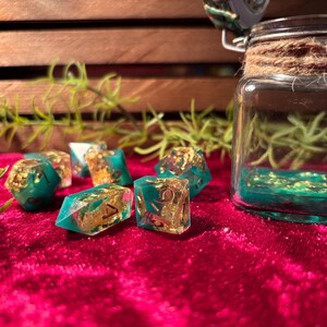 Pode incluir: Um conjunto de dados de resina verde-azulado e dourada com inclus&otilde;es de folha de ouro, exibidos em uma superf&iacute;cie de veludo vermelho. Um frasco de vidro com l&iacute;quido verde-azulado e folha de ouro ao fundo.