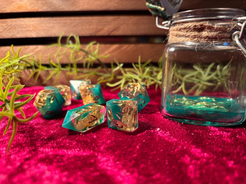 Pode incluir: Um conjunto de dados de resina verde-azulado e dourada com inclus&otilde;es de folha de ouro, exibidos em uma superf&iacute;cie de veludo vermelho. Um frasco de vidro com l&iacute;quido verde-azulado e folha de ouro ao fundo.