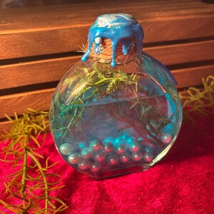 May include: A clear, round glass bottle with a blue wax seal and a cork stopper. The bottle contains blue liquid, green sprigs, and blue pearl-like beads. The bottle sits on a red velvet surface.