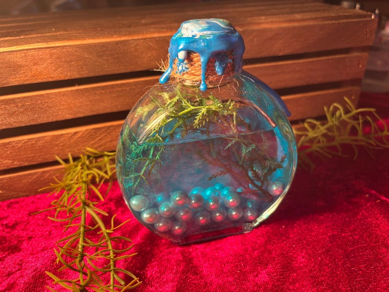 May include: A clear, round glass bottle with a blue wax seal and a cork stopper. The bottle contains blue liquid, green sprigs, and blue pearl-like beads. The bottle sits on a red velvet surface.