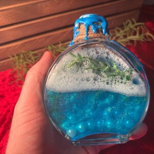 May include: A clear, round glass bottle filled with blue liquid, white foam, and green sprigs. The bottle is sealed with a cork and blue paint. The bottle contains small, light blue pearls.