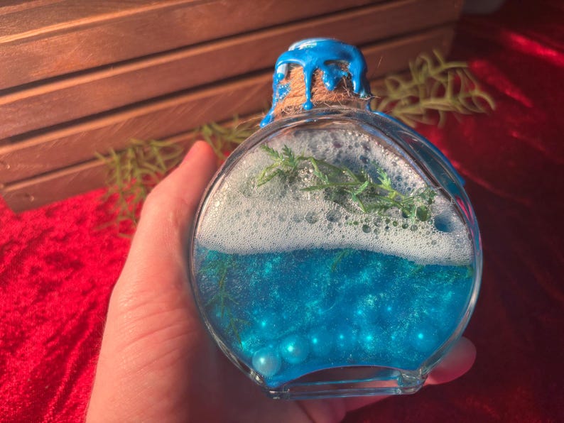 May include: A clear, round glass bottle filled with blue liquid, white foam, and green sprigs. The bottle is sealed with a cork and blue paint. The bottle contains small, light blue pearls.
