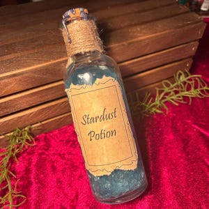 May include: A clear glass bottle filled with blue liquid labeled "Stardust Potion." The bottle is sealed with a cork and twine, and sits on a red velvet surface. The background includes a wooden box.