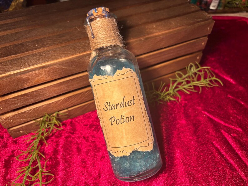 May include: A clear glass bottle filled with blue liquid labeled "Stardust Potion." The bottle is sealed with a cork and twine, and sits on a red velvet surface. The background includes a wooden box.