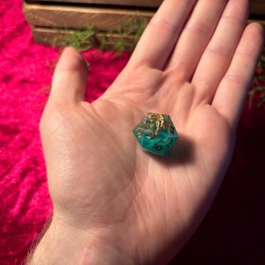 Pode incluir: Um dado D20 de resina turquesa e dourada, com o n&uacute;mero 10 vis&iacute;vel. O dado &eacute; segurado em uma m&atilde;o aberta contra um fundo de veludo vermelho. A parte superior do dado &eacute; dourada e as laterais s&atilde;o turquesa.