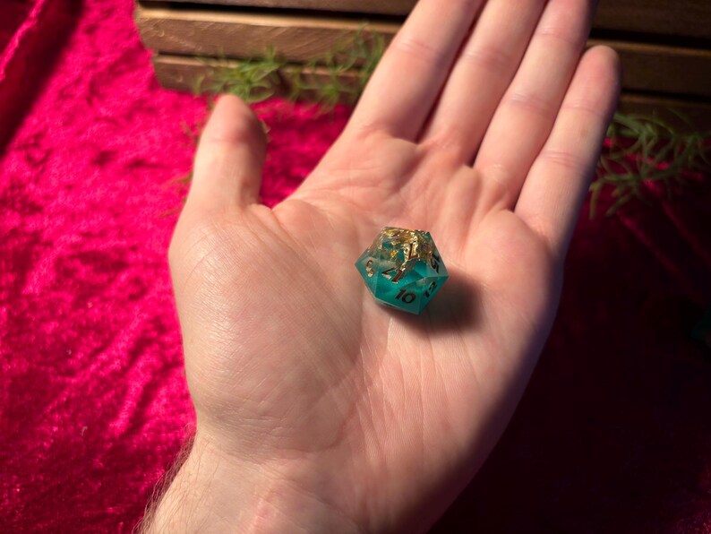 Pode incluir: Um dado D20 de resina turquesa e dourada, com o n&uacute;mero 10 vis&iacute;vel. O dado &eacute; segurado em uma m&atilde;o aberta contra um fundo de veludo vermelho. A parte superior do dado &eacute; dourada e as laterais s&atilde;o turquesa.