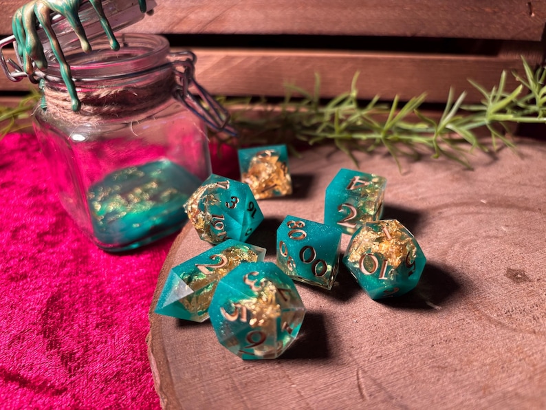 Pode incluir: Um conjunto de dados de resina azul-turquesa com inclus&otilde;es de folha de ouro, espalhados sobre uma superf&iacute;cie de madeira. Um frasco de vidro com resina azul-turquesa e folha de ouro est&aacute; no fundo. Os dados t&ecirc;m v&aacute;rias formas.