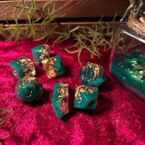 Pode incluir: Um conjunto de dados de resina verde-azulado e dourada com numera&ccedil;&atilde;o dourada, espalhados sobre uma superf&iacute;cie de veludo vermelho escuro. Um frasco de vidro com resina verde-azulado e flocos de ouro est&aacute; no fundo, juntamente com raminhos de vegeta&ccedil;&atilde;o.