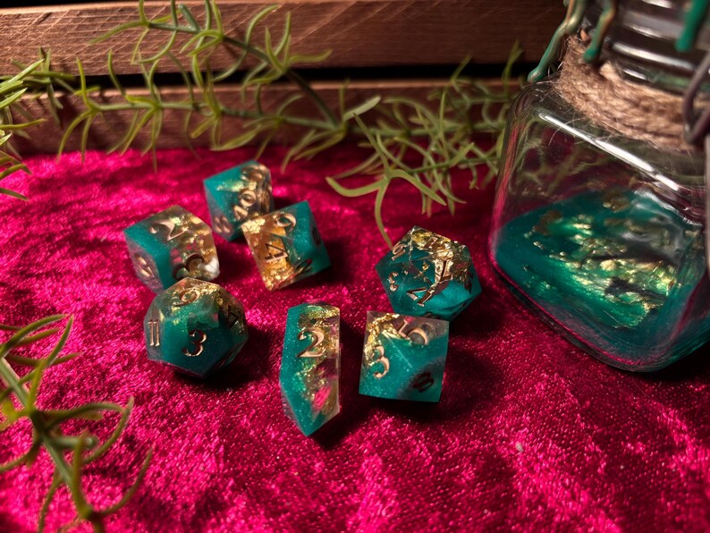 Pode incluir: Um conjunto de dados de resina verde-azulado e dourada com numera&ccedil;&atilde;o dourada, espalhados sobre uma superf&iacute;cie de veludo vermelho escuro. Um frasco de vidro com resina verde-azulado e flocos de ouro est&aacute; no fundo, juntamente com raminhos de vegeta&ccedil;&atilde;o.