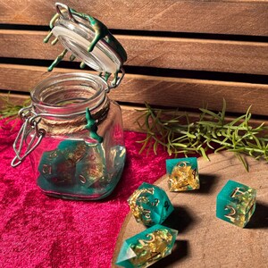 Pode incluir: Um frasco de vidro com tampa articulada, cheio de dados turquesa e dourados. V&aacute;rios dados est&atilde;o espalhados ao redor do frasco. O frasco e os dados est&atilde;o em uma superf&iacute;cie rosa, com uma caixa de madeira ao fundo.