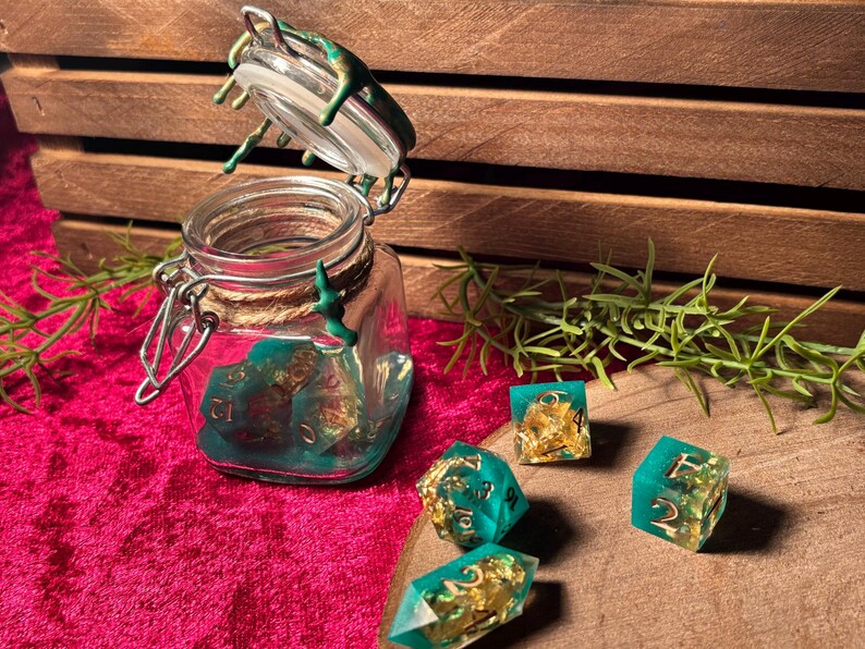 Pode incluir: Um frasco de vidro com tampa articulada, cheio de dados turquesa e dourados. V&aacute;rios dados est&atilde;o espalhados ao redor do frasco. O frasco e os dados est&atilde;o em uma superf&iacute;cie rosa, com uma caixa de madeira ao fundo.