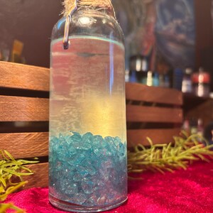May include: A clear glass bottle filled with a light-colored liquid and blue crystals. The bottle is sealed with a cork and twine. The background includes wooden crates and a red velvet surface.