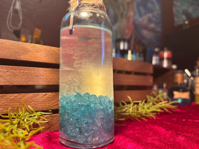 May include: A clear glass bottle filled with a light-colored liquid and blue crystals. The bottle is sealed with a cork and twine. The background includes wooden crates and a red velvet surface.