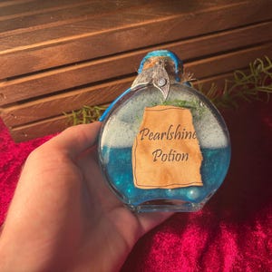 May include: A round glass bottle filled with blue liquid and white foam, labelled "Pearlshine Potion." The bottle has a decorative top and is held against a red velvet background.