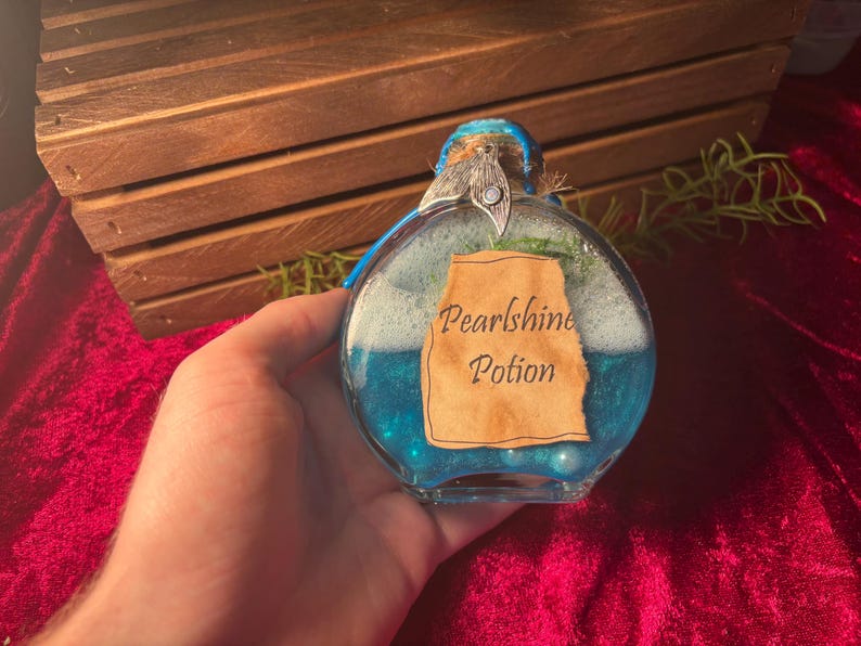 May include: A round glass bottle filled with blue liquid and white foam, labelled "Pearlshine Potion." The bottle has a decorative top and is held against a red velvet background.