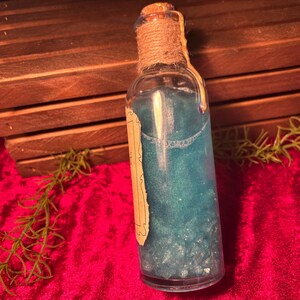 May include: A clear glass bottle filled with a shimmering blue liquid. The bottle is sealed with a cork and twine, and has a vintage-style label. The background includes a wooden crate and red fabric.