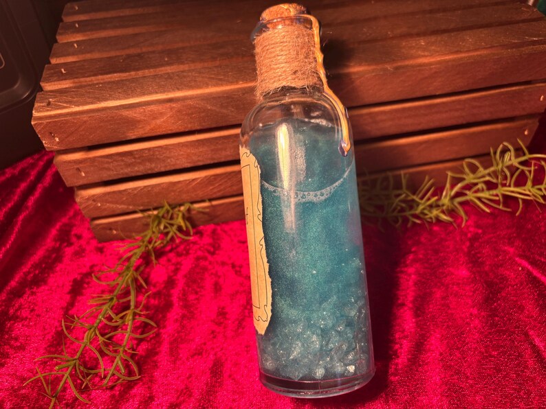 May include: A clear glass bottle filled with a shimmering blue liquid. The bottle is sealed with a cork and twine, and has a vintage-style label. The background includes a wooden crate and red fabric.