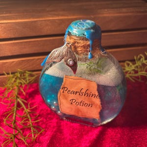May include: A glass bottle filled with blue liquid and small pearls, labelled "Pearlshine Potion." The bottle has a blue-painted top, a cork stopper, and a decorative silver charm. The background includes a wooden crate and red fabric.