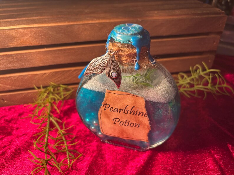 May include: A glass bottle filled with blue liquid and small pearls, labelled "Pearlshine Potion." The bottle has a blue-painted top, a cork stopper, and a decorative silver charm. The background includes a wooden crate and red fabric.