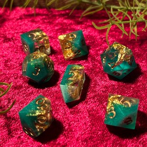 Pode incluir: Um conjunto de sete dados de resina verde-azulado e dourada. Os dados t&ecirc;m detalhes em folha de ouro e est&atilde;o colocados sobre uma superf&iacute;cie de veludo vermelho escuro. Raminhos verdes no fundo.