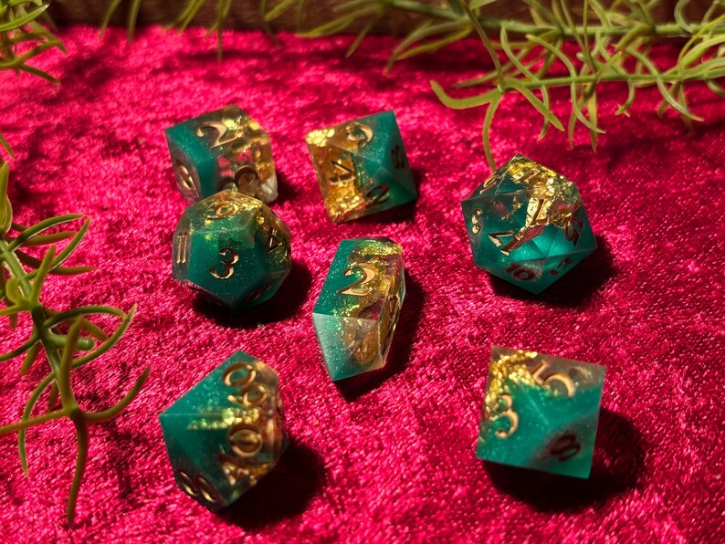 Pode incluir: Um conjunto de sete dados de resina verde-azulado e dourada. Os dados t&ecirc;m detalhes em folha de ouro e est&atilde;o colocados sobre uma superf&iacute;cie de veludo vermelho escuro. Raminhos verdes no fundo.