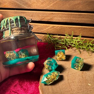 Pode incluir: Um frasco de vidro com fecho met&aacute;lico, cheio de l&iacute;quido e dados. O frasco &eacute; decorado com pingos verdes e uma base turquesa. V&aacute;rios dados turquesa e dourados est&atilde;o espalhados sobre uma superf&iacute;cie de madeira, sugerindo um tema de jogo de tabuleiro.