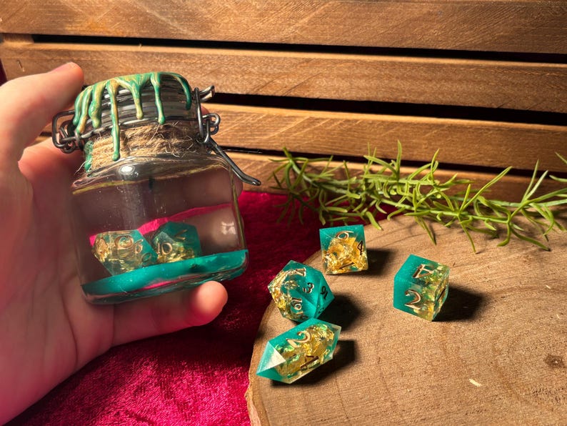 Pode incluir: Um frasco de vidro com fecho met&aacute;lico, cheio de l&iacute;quido e dados. O frasco &eacute; decorado com pingos verdes e uma base turquesa. V&aacute;rios dados turquesa e dourados est&atilde;o espalhados sobre uma superf&iacute;cie de madeira, sugerindo um tema de jogo de tabuleiro.