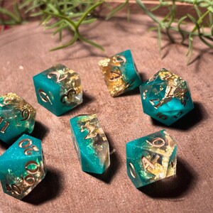 Pode incluir: Conjunto de sete dados poli&eacute;dricos com base em resina turquesa e inclus&otilde;es de folha de ouro. Os n&uacute;meros s&atilde;o em fonte met&aacute;lica dourada. Os dados est&atilde;o dispostos sobre uma superf&iacute;cie de madeira.