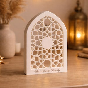 May include: Cream-coloured decorative lantern featuring a geometric design and the text "The Ahmed Family". The lantern sits on a wooden surface. A vase with dried plants and a decorative lantern are in the background.