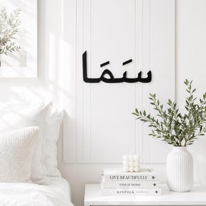 Op de afbeelding: Een rechthoekig wit wandpaneel met verticale groeven en zwarte Arabische kalligrafie in het midden. Het kunstwerk hangt boven een wit nachtkastje met boeken, een kleine witte kaars en een witte vaas met groene bladeren.