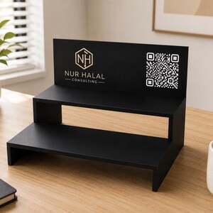 May include: A black two-tiered display stand with the text "NUR HALAL CONSULTING" and a QR code. The stand is made of wood and is on a light brown wooden surface. A small plant in a white pot is to the left of the stand.