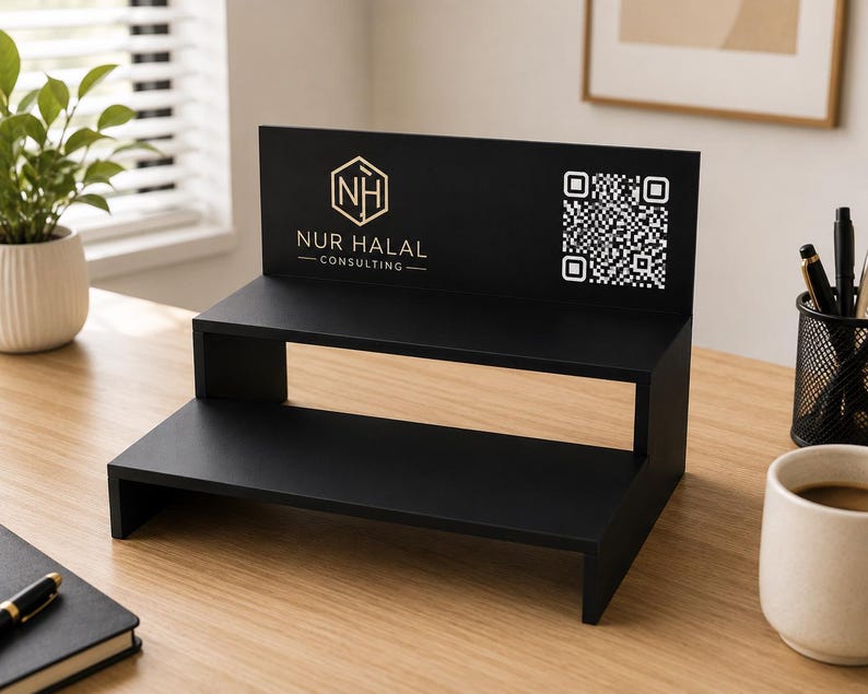 May include: A black two-tiered display stand with the text "NUR HALAL CONSULTING" and a QR code. The stand is made of wood and is on a light brown wooden surface. A small plant in a white pot is to the left of the stand.