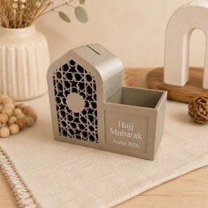 May include: A gray decorative money box with a geometric design and the words "Hajj Mubarak Aisha 2026". The money box has a slot for inserting money and a small compartment. The box is made of a light gray material.