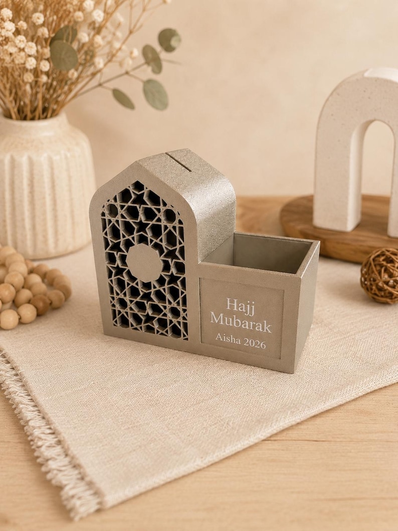 May include: A gray decorative money box with a geometric design and the words "Hajj Mubarak Aisha 2026". The money box has a slot for inserting money and a small compartment. The box is made of a light gray material.