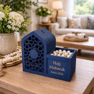 May include: A navy blue decorative box with a house-shaped design and geometric cutouts. The box has a compartment filled with pearls and text that reads "Hajj Mubarak Aisha 2026".