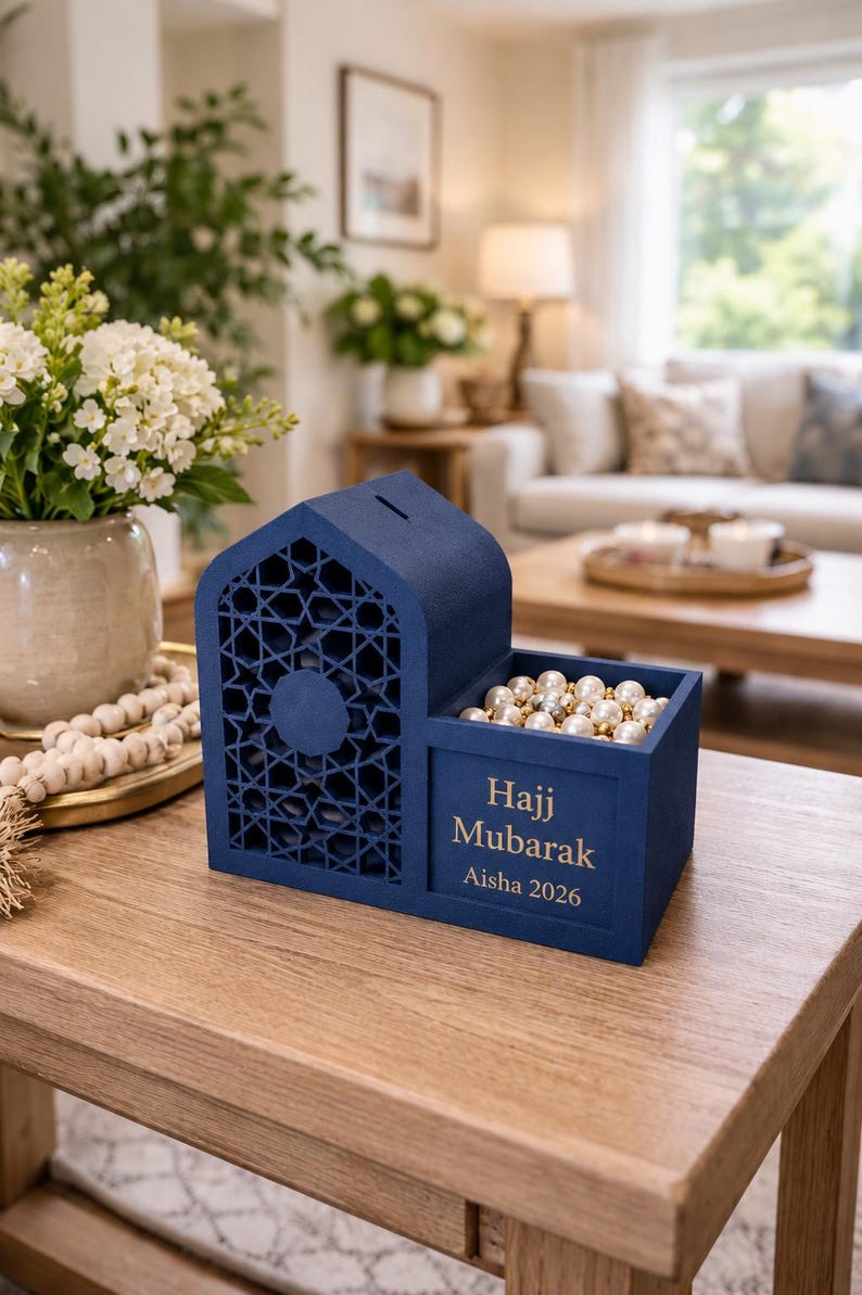 May include: A navy blue decorative box with a house-shaped design and geometric cutouts. The box has a compartment filled with pearls and text that reads "Hajj Mubarak Aisha 2026".