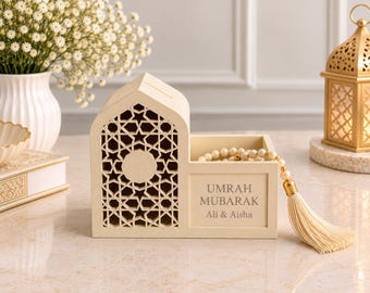 May include: A cream-colored wooden box with an arched, laser-cut geometric star pattern on one side and an open compartment on the other. The front features the text "UMRAH MUBARAK" and "Ali & Aisha" with a cream-colored beaded tassel attached.