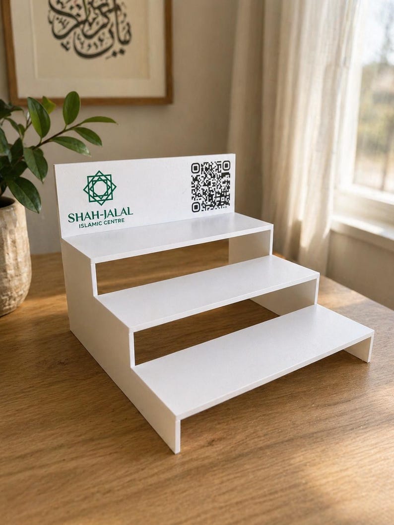 May include: A white, three-tiered display stand with the text "SHAH-JALAL ISLAMIC CENTRE" and a geometric logo in green. A QR code is printed on the top tier. The stand is made of a solid material and is designed to showcase items.