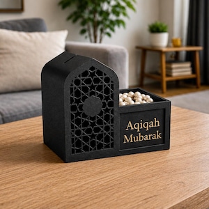 May include: A black wooden money box with a decorative geometric design and the words "Aqiqah Mubarak" in gold lettering. The box is filled with small, round, white objects and sits on a wooden table.