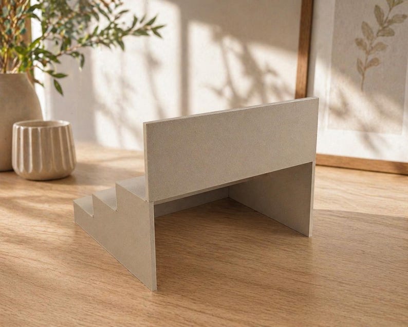 May include: A light beige, three-step display stand with a back panel. The stand is made of a solid material and is positioned on a wooden surface. The design is minimalist and modern, suitable for showcasing small items.