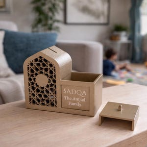 May include: A tan-colored decorative box with a geometric design and the text "SADQA The Amjad Family." The box has a slot on top and a separate lid. The box is on a wooden table.