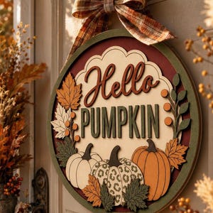 Może przedstawiać: Okrągła drewniana tabliczka na drzwi z napisem Hello PUMPKIN wykonanym z warstwowych liter. Wzór zawiera trzy dynie, w tym jedną w panterkę, otoczone jesiennymi liśćmi oraz kokardą z materiału w kratę umieszczoną na górze.
