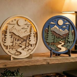 May include: Two circular layered wooden art pieces featuring mountain landscapes with pine trees, a winding river, and a moon. One piece is monochromatic light wood, while the other features blue, green, and brown tones with integrated lighting.